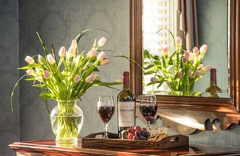 A dresser or side table with vase and pink tulip bulbs, next to a tray of red wine, with a mirror in the background, tulips visible in mirror.